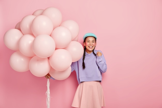 Glückliches asiatisches Mädchen ballt die Faust vor Freude, kann es kaum erwarten, einen besonderen Moment zu erleben, bekommt Glückwünsche von Freunden zu ihrem Geburtstag, trägt einen großen Haufen Luftballons, gekleidet in fahionable Kleidung