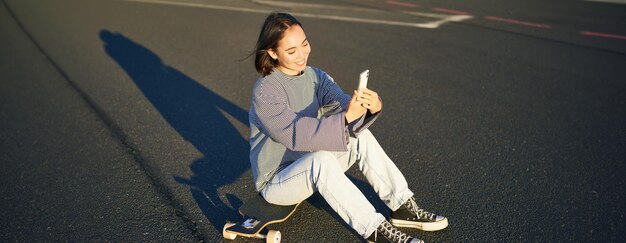 Kostenloses Foto glückliches asiatisches mädchen sitzt auf einem skateboard, macht ein selfie mit einem longboard, macht süße gesichter an einem sonnigen tag im freien.