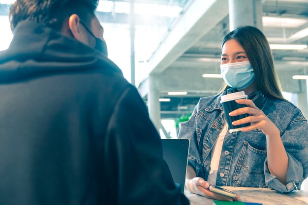 Glückliches asiatisches Paar trägt Gesichtsmasken-Virenschutz in einem Café und genießt ein gutes Gespräch Junger Mann und Frau in einem Restaurant, die sich den Touchscreen-Computer ansehen, lachen, lächeln zusammen