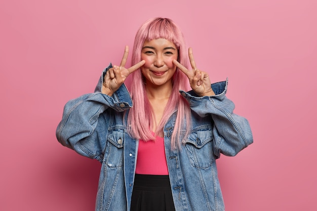 Glückliches asiatisches Teenager-Mädchen mit langen rosa Haaren, gekleidet in Jeanskleidung, zeigt Friedens- oder Siegeszeichen, versicherter Tag sei großartig, hat Spaß