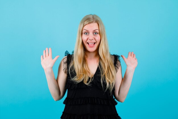 Glückliches blondes Mädchen schaut in die Kamera, indem sie ihre Handvoll auf blauem Hintergrund erhebt