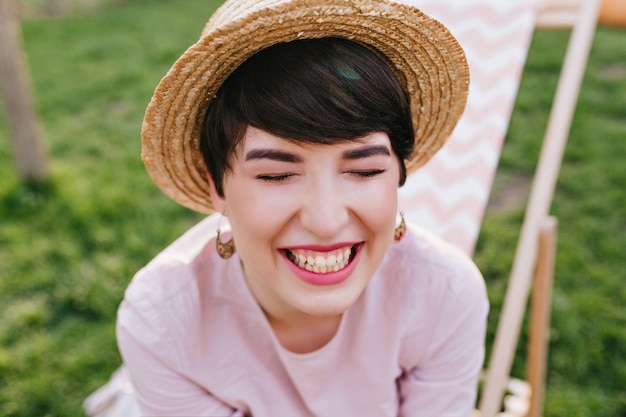 Glückliches brünettes mädchen im trendigen sommerhut und in den niedlichen ohrringen, die mit geschlossenen augen lachen