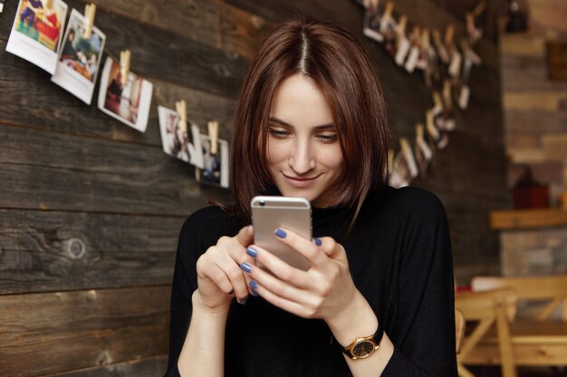 Glückliches brünettes Mädchen in der schwarzen Kleidung unter Verwendung der freien drahtlosen Internetverbindung auf Smartphone, während Ruhe im Restaurant mit gemütlichem Interieur und Bildern, die an Holzwand hängen