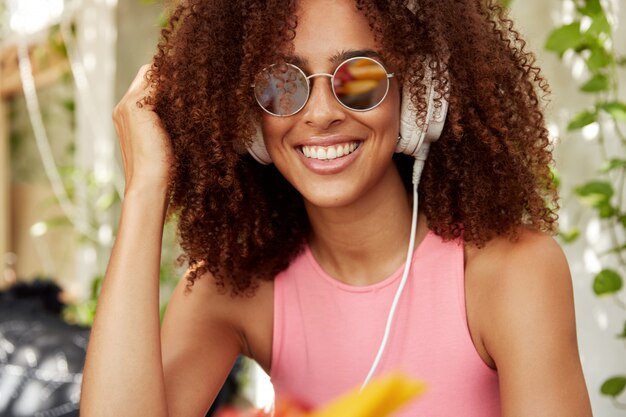 Glückliches dunkelhäutiges lockiges weibliches Model in trendigen Farben, genießt Musik in großen Kopfhörern, hat ein strahlendes Lächeln oder hört Radio. Schöne afrikanische Frau hört Lieblingssendung mit Kopfhörern