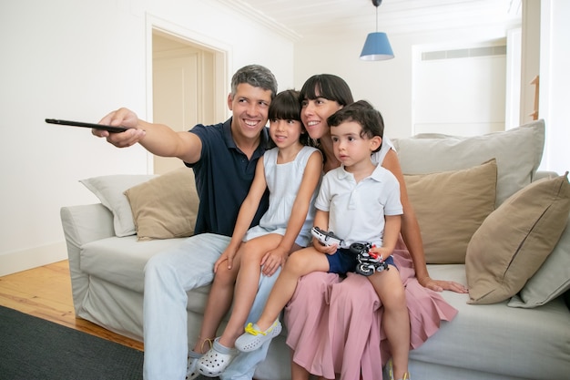 Glückliches Elternpaar mit zwei Kindern, die fernsehen, auf der Couch im Wohnzimmer sitzen und Fernbedienung verwenden.