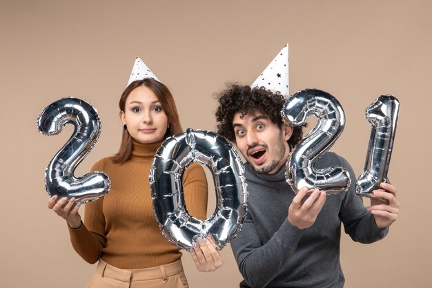 Glückliches energisches junges Paar tragen Neujahrshut posiert für Kamera Mädchen zeigt und und Kerl mit und auf Grau