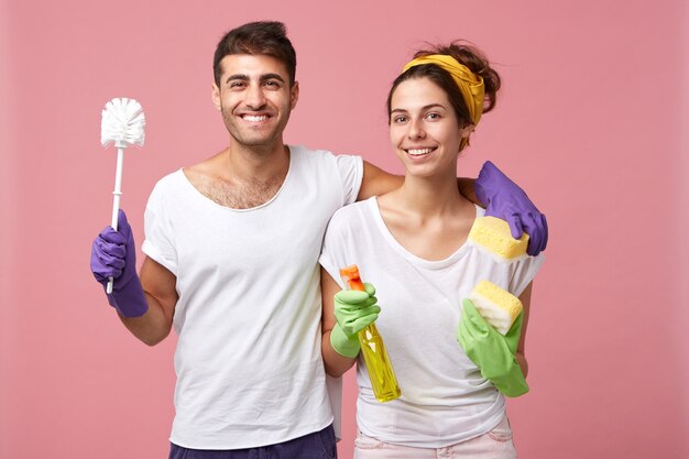 Glückliches europäisches Paar mit Schwamm, Waschmittel und Bürste, die gute Laune vor dem Frühjahrsputz umarmen, gute Beziehungen haben und Hausarbeit zusammen machen. Hausputz- und Teamwork-Konzept
