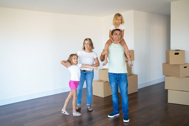 Glückliches Familienpaar und zwei Kinder, die über ihre neue Wohnung schauen und in einem leeren Raum mit Stapeln von Kisten stehen