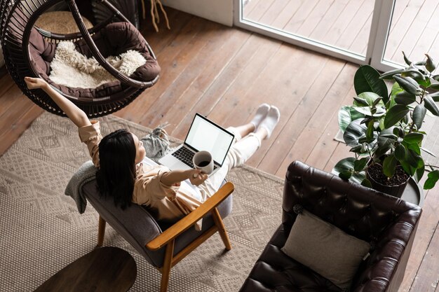 Glückliches freies asiatisches Mädchen, das zu Hause mit Laptop und Kaffeetasse sitzt und die Hände hochhebt