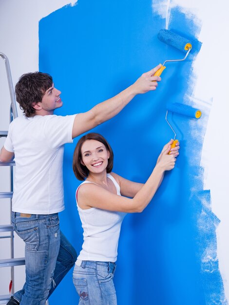 Glückliches fröhliches Paar mit Rollen, die die Wand malen - drinnen