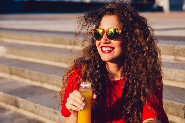 Glückliches gelocktes Mädchen in der Sonnenbrille hält eine Flasche mit dem frischen Saft und genießt einen sonnigen Tag
