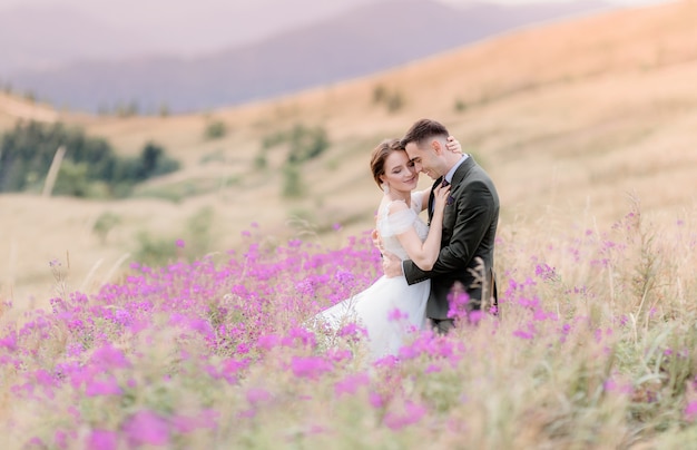 Glückliches hochzeitspaar sitzt auf dem hügel der wiese, umgeben von rosa blumen