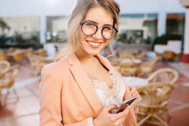 Glückliches hübsches mädchen mit grauem smartphone in der hand, lächelnd, student, geschäftsdame. straßencafé, terrasse. tragen sie eine stilvolle brille, eine rosa jacke und eine beige spitzenbluse.