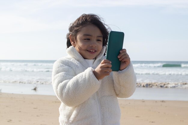 Glückliches japanisches Mädchen mit Handy am Strand. Dunkelhaariges Kind, das ein Gerät hält und auf den Bildschirm schaut. Kindheit, Technologiekonzept
