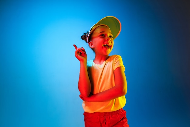 Glückliches jugendlich mädchen, das steht, lächelt und über trendigen blauen neonraum zeigt. schönes weibliches porträt. junge befriedigen mädchen