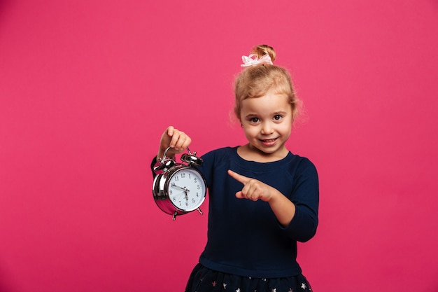 Glückliches junges blondes Mädchen, das Wecker hält und auf ihn beim Betrachten der Kamera über rosa Wand zeigt