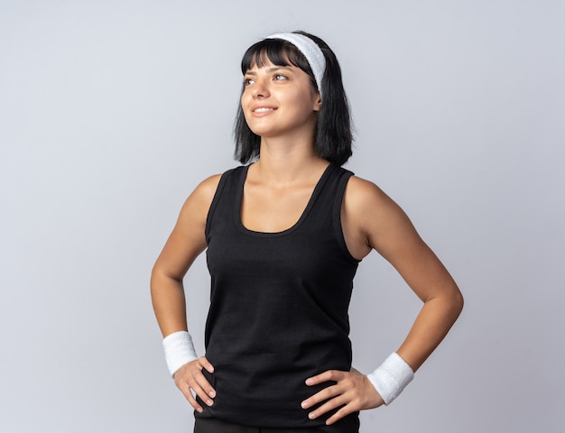 Glückliches junges Fitness-Mädchen mit Stirnband, das fröhlich lächelnd auf weißem Hintergrund aufschaut