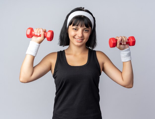 Glückliches junges Fitness-Mädchen mit Stirnband, das Hanteln hält und Übungen macht, die selbstbewusst lächelnd aussehen