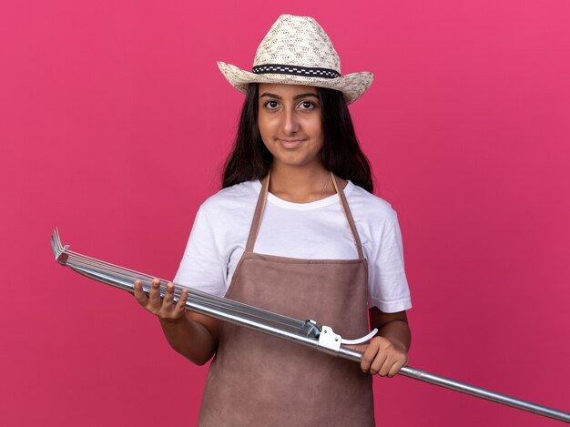 Glückliches junges Gärtnermädchen in der Schürze und im Sommerhut, die Rechen mit Lächeln auf Gesicht halten, das über rosa Wand steht