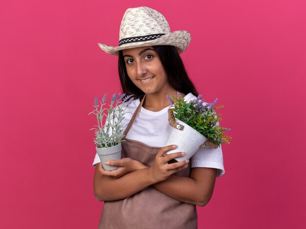 Glückliches junges Gärtnermädchen in der Schürze und im Sommerhut, die Topfpflanzen lächelnd fröhlich stehend über rosa Wand halten