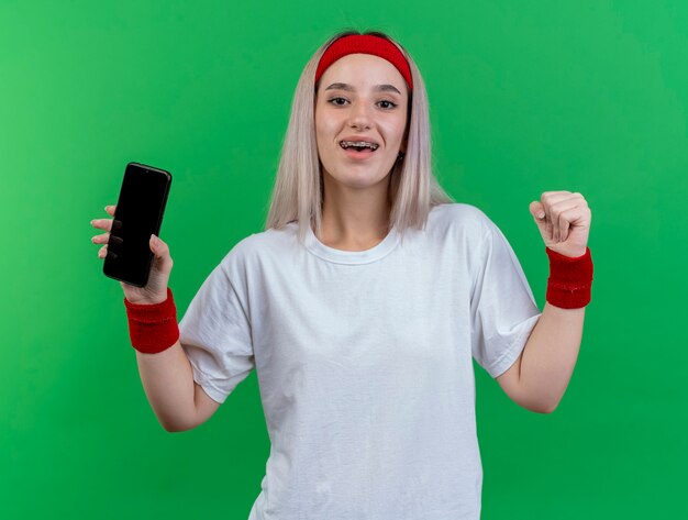 Glückliches junges kaukasisches sportliches Mädchen mit Hosenträgern, das Stirnband und Armbänder trägt, hält die Faust und hält das Telefon