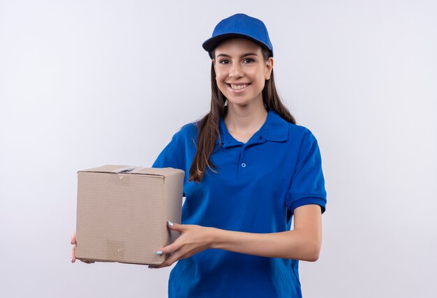 Glückliches junges Liefermädchen in der blauen Uniform und in der Kappe, die Boxpaket hält, das zuversichtlich lächelt