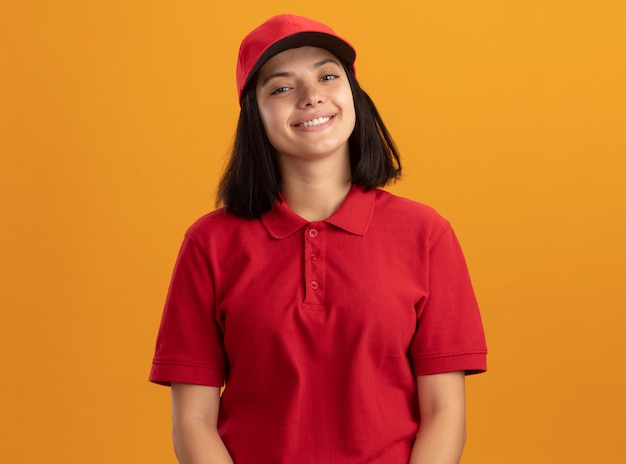 Glückliches junges Liefermädchen in der roten Uniform und in der Kappe, die fröhlich über orange Wand stehend lächeln