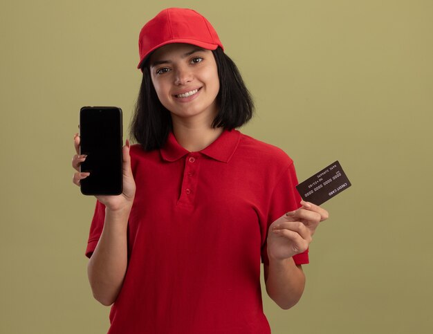 Glückliches junges Liefermädchen in der roten Uniform und in der Kappe, die Smartphine und Kreditkarte zeigt, die fröhlich stehen über grüner Wand lächeln