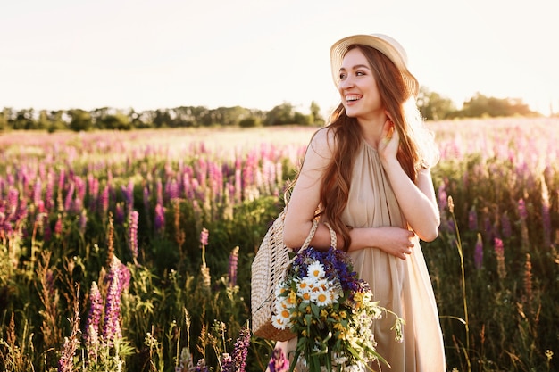 Glückliches junges Mädchen, das auf dem Blumengebiet bei Sonnenuntergang geht. Strohhut und Tasche voller Blumen tragen.