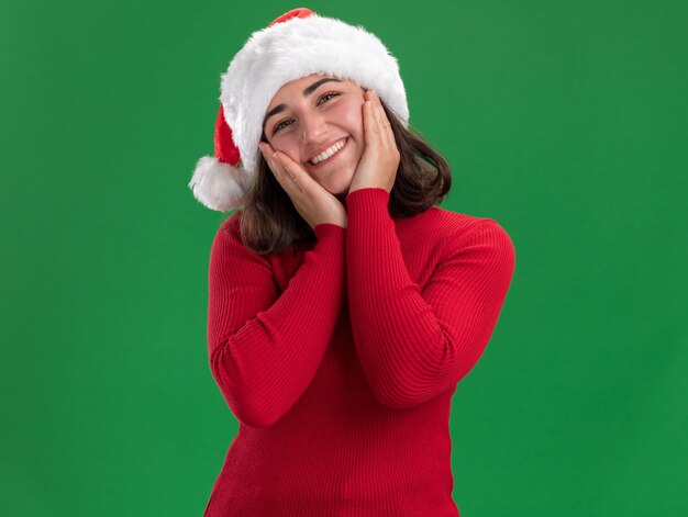 Kostenloses Foto glückliches junges mädchen im roten pullover und im weihnachtsmannhut lächelnd breit stehend über grüner wand