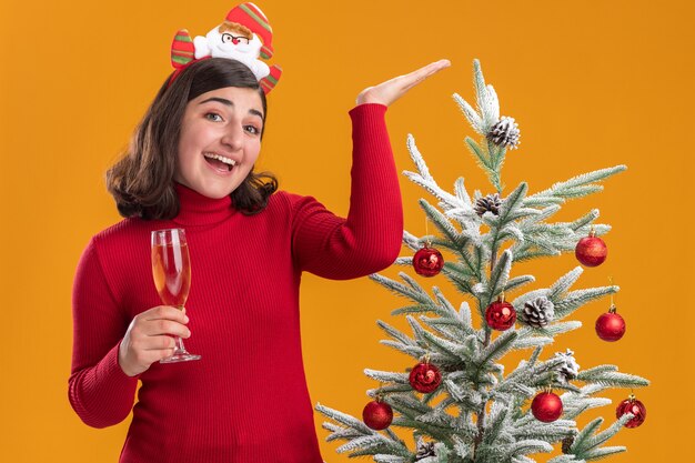 Glückliches junges Mädchen im Weihnachtspullover, der lustiges Stirnband hält, das Glas Champagner hält, der fröhlich mit dem Arm angehoben trägt, der neben einem Weihnachtsbaum über orange Wand steht