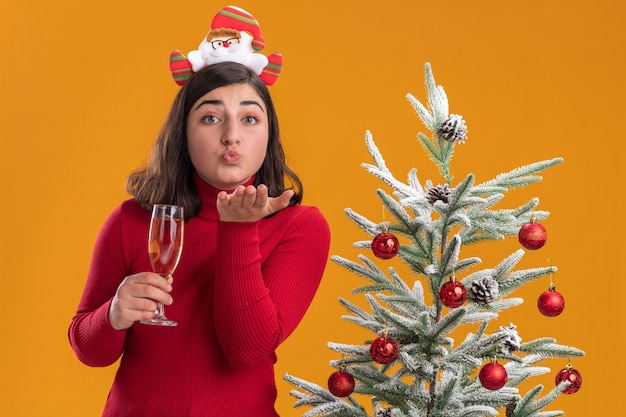 Glückliches junges Mädchen im Weihnachtspullover, der lustiges Stirnband trägt, das Glas Champagner hält und einen Kuss neben einem Weihnachtsbaum über orange Hintergrund bläst