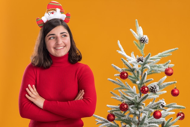 Glückliches junges Mädchen im Weihnachtspullover, der lustiges Stirnband trägt, das mit Lächeln auf Gesicht neben einem Weihnachtsbaum über orange Hintergrund beiseite schaut