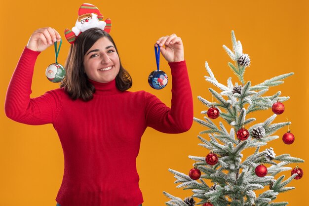 Glückliches junges Mädchen im Weihnachtspullover, der lustiges Stirnband trägt, das Weihnachtskugeln mit Lächeln auf Gesicht hält, das neben einem Weihnachtsbaum über orange Wand steht