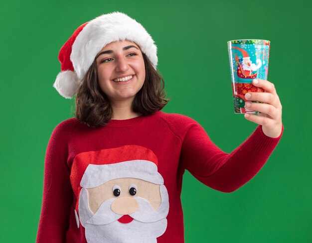 Glückliches junges Mädchen im Weihnachtspullover, der Weihnachtsmannmütze hält, der bunte Pappbecher hält, der es mit Lächeln auf Gesicht betrachtet, das über grüner Wand steht