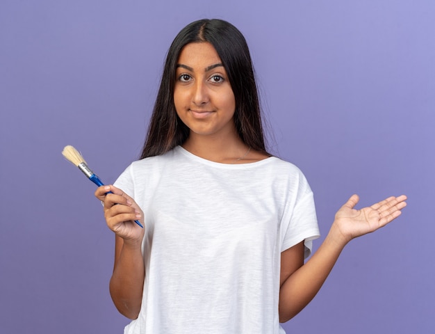 Glückliches junges Mädchen im weißen T-Shirt mit Pinsel in die Kamera schaut mit einem Lächeln auf dem Gesicht auf blauem Hintergrund