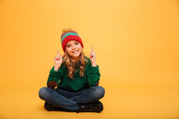 Glückliches junges Mädchen in der Strickjacke und in Hut, die auf dem Boden beim die Kamera oben zeigen und betrachten über Orange sitzen