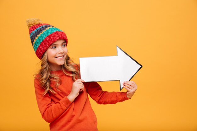 Glückliches junges Mädchen in der Strickjacke und in Hut, die mit Papierpfeil weg über Orange schauen und zeigen