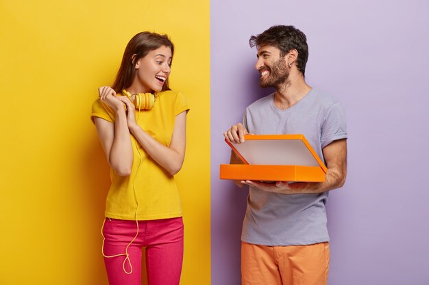 Glückliches junges männliches Modell öffnet Schachtel, überrascht Freundin am Geburtstag, zeigt etwas im Paket