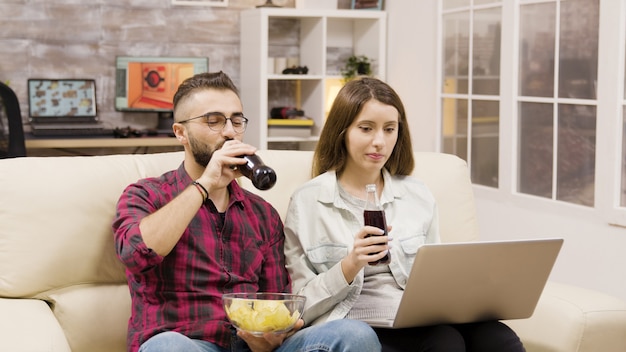 Glückliches junges Paar beim Online-Shopping auf dem Laptop. Paar sitzt auf der Couch, trinkt Soda und isst Chips.