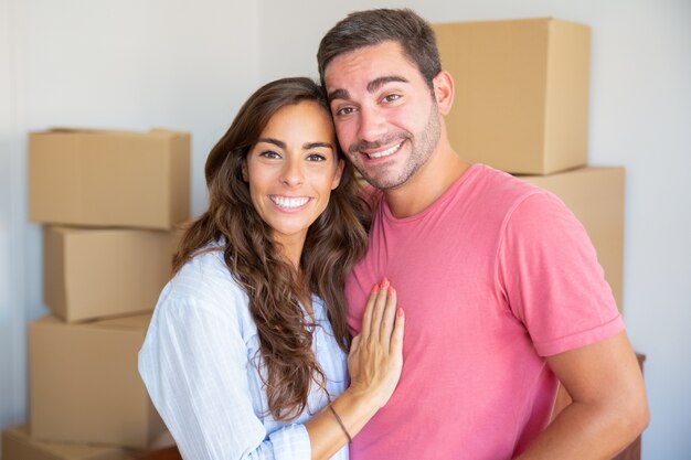 Glückliches junges Paar, das genießt, in neue Wohnung zu ziehen, zwischen Kartonschachteln stehend, Umarmung und Kamera betrachtend