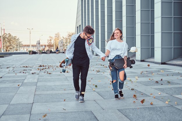 Glückliches junges Paar hält Händchen und geht bei windigem Wetter mit Skateboards auf einer modernen Straße spazieren.