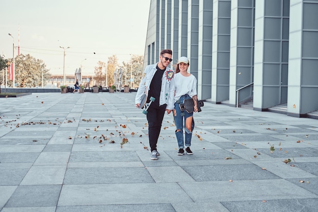 Glückliches junges Paar hält Händchen und geht bei windigem Wetter mit Skateboards auf einer modernen Straße spazieren.