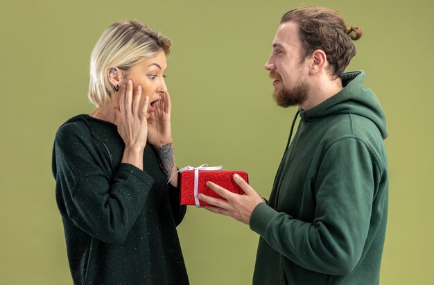 glückliches junges Paar im Freizeitkleidungsmann mit Geschenk für seine reizende lächelnde und überraschte Freundin, die Valentinstag feiert, der über grüner Wand steht