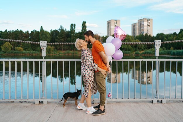 Glückliches junges Paar umarmt und küsst auf Brücke, die rosa Luftballons und Hund mit Stadt am Horizont hält