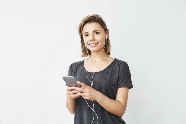 Glückliches junges schönes Mädchen in den Kopfhörern, die Telefon lächelnd halten.