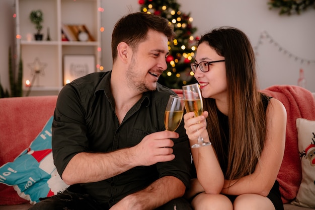 Glückliches junges und schönes Paar mit Gläsern Champagner sitzt auf der Couch und feiert zusammen Weihnachten im weihnachtlich dekorierten Raum mit Weihnachtsbaum im Hintergrund