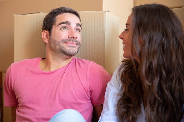 Glückliches junges verliebtes Paar, das auf Boden nahe Haufen von Kartonschachteln sitzt und genießt, in neue Wohnung einziehen