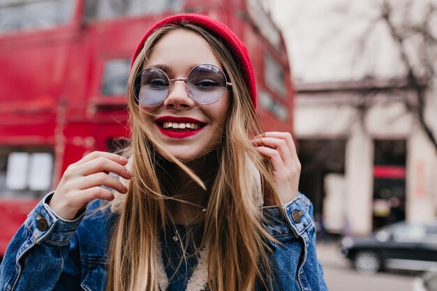 Glückliches kaukasisches Mädchen in der Jeansjacke, die auf Unschärfestraße aufwirft. Außenaufnahme der bezaubernden weißen Dame in der stilvollen blauen Brille.