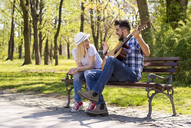 Glückliches kaukasisches Paar, das auf einer Parkbank sitzt, mit dem Mann, der bei Tageslicht Gitarre spielt